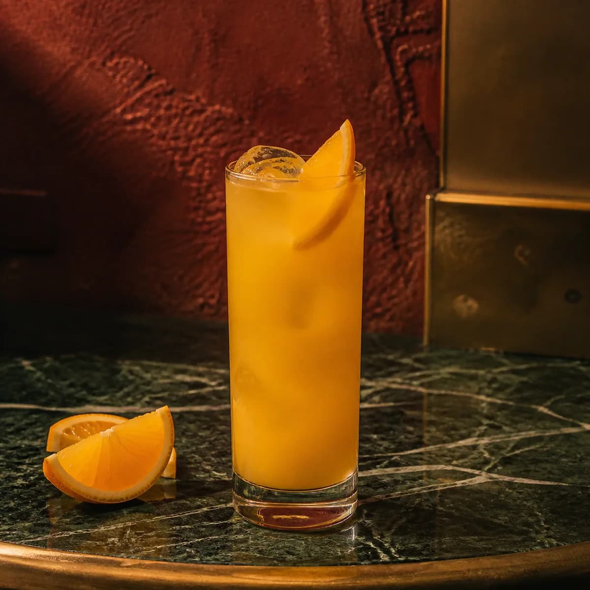 Fuzzy Navel in a tall straight-sided highball glass styled in a Milan aperitivo bar with oxblood hand-painted plaster, asymmetric aged brass and dark green marble.