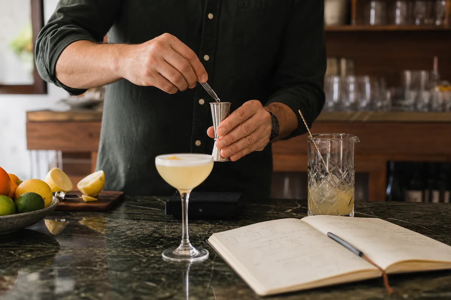 A recipe tester measuring a small correction beside a cocktail, mixing glass and open notebook.