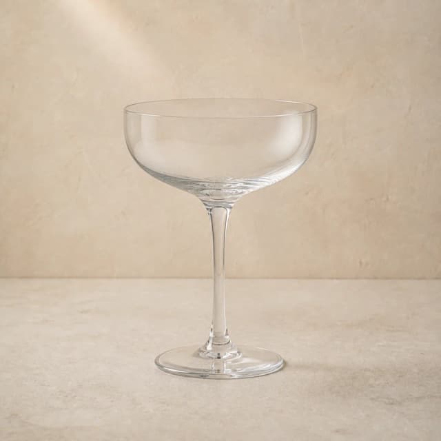 An empty clear coupe glass with a shallow rounded bowl and slender stem.