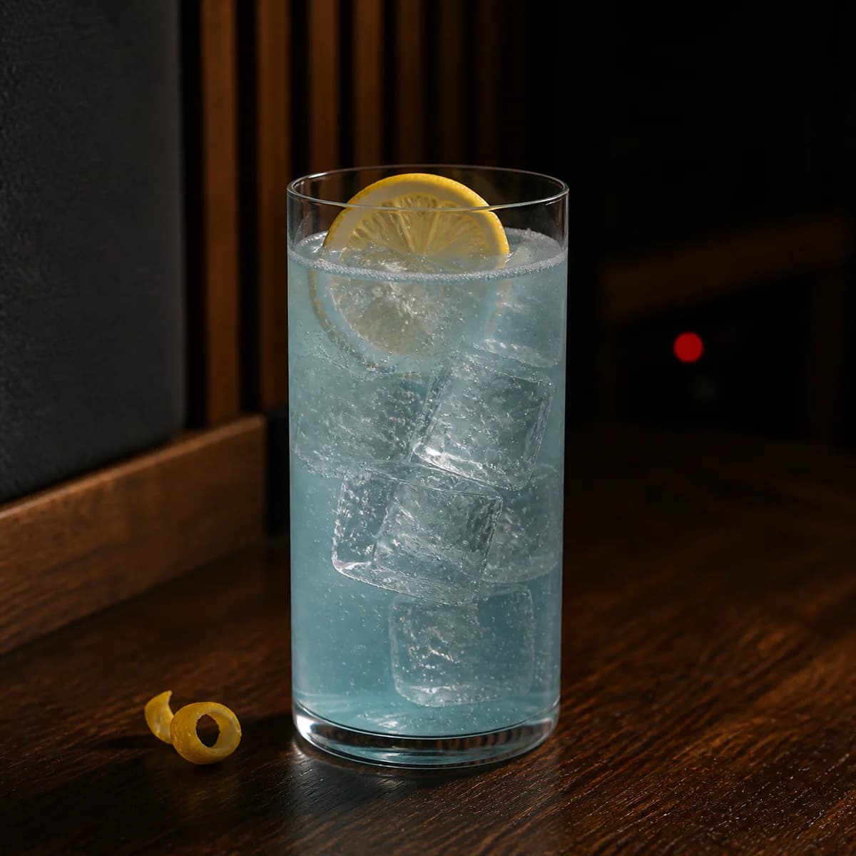 Blue Lagoon in a tall straight-sided highball glass styled in a small listening bar with acoustic walnut, dark felt and a dim red indicator in the distance.