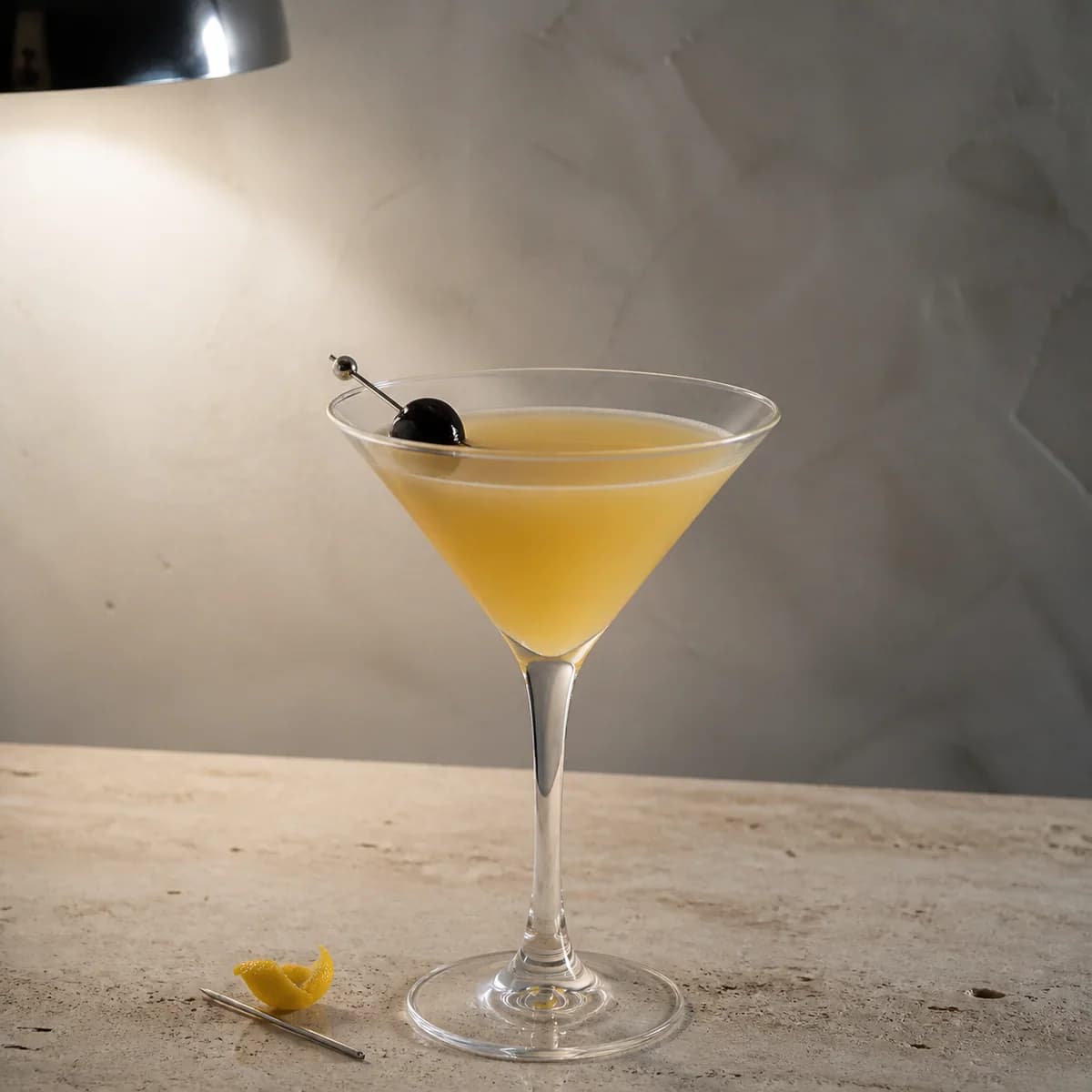 French Martini in a sharp V-shaped martini glass styled in a quiet minimalist bar with chalky gray plaster, pale travertine and one off-centre chrome light.