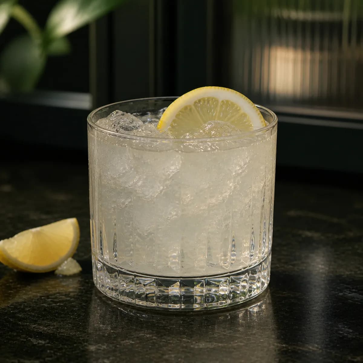 Gin Fix in a heavy low rocks glass styled in a restrained botanical bar with dark green painted wood, ribbed glass and a few out-of-focus leaves.