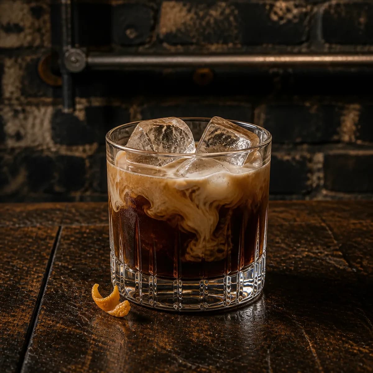White Russian in a heavy low rocks glass styled in a lived-in industrial loft bar with charcoal brick, scratched walnut and aged metal details.